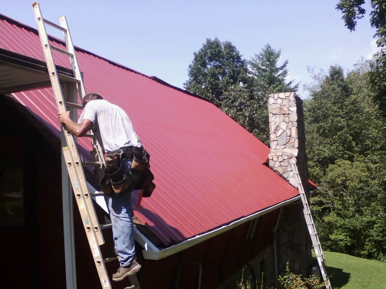 Roofer on a ladder beside a red metal roof and stone chimney in Linwood